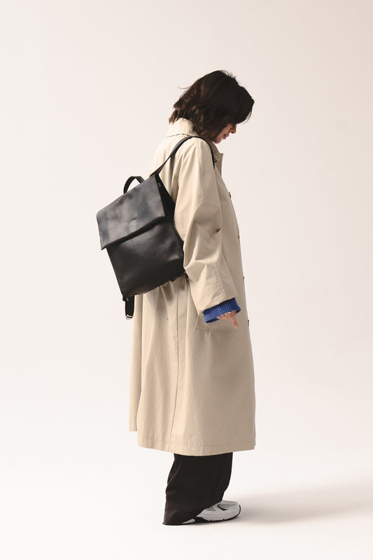 Czarny skórzany plecak Fi marki Taksuba noszony na plecach przez kobietę w beżowym trenczu