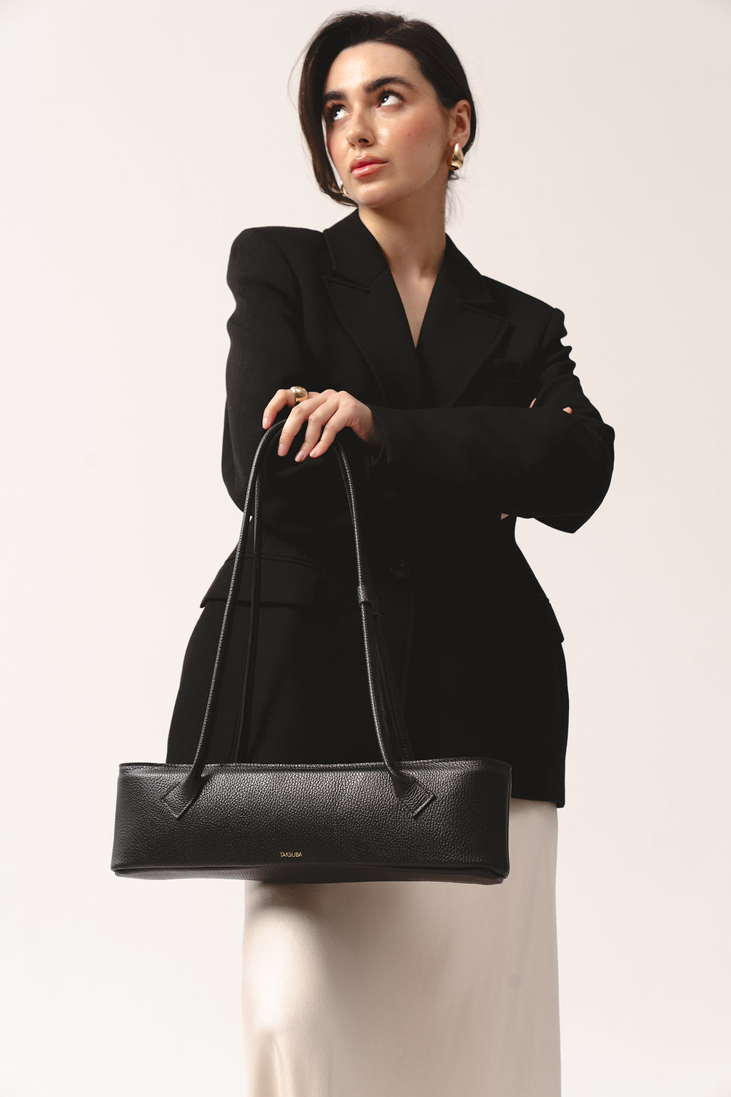 Woman in a tailored black blazer and white skirt holding a black pebbled leather TAKSUBA Teckelete handbag with long handles.