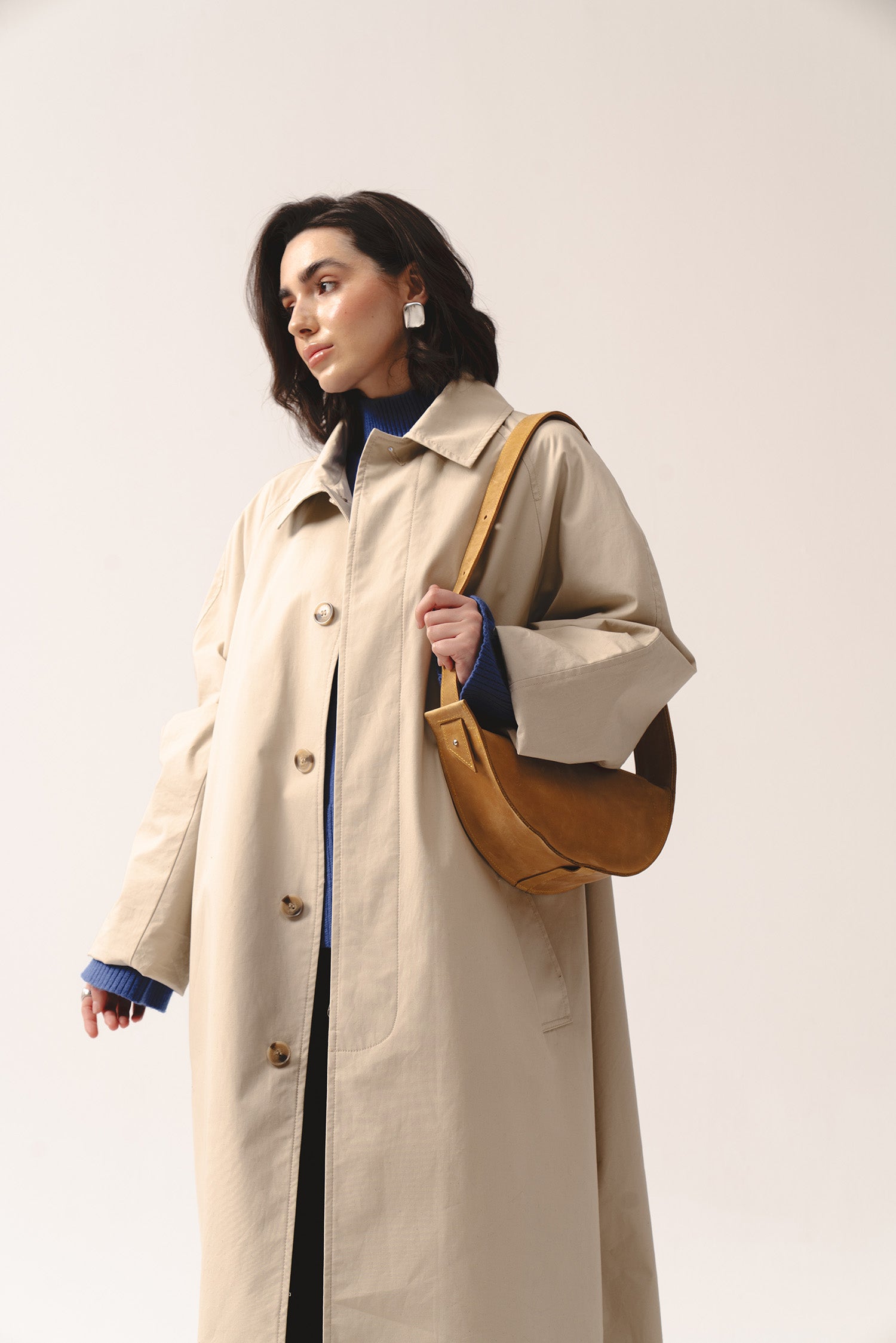 Kobieta w beżowym płaszczu i niebieskim swetrze z żółtą skórzaną torebką półksiężyc Eclipse M przewieszoną przez ramię.