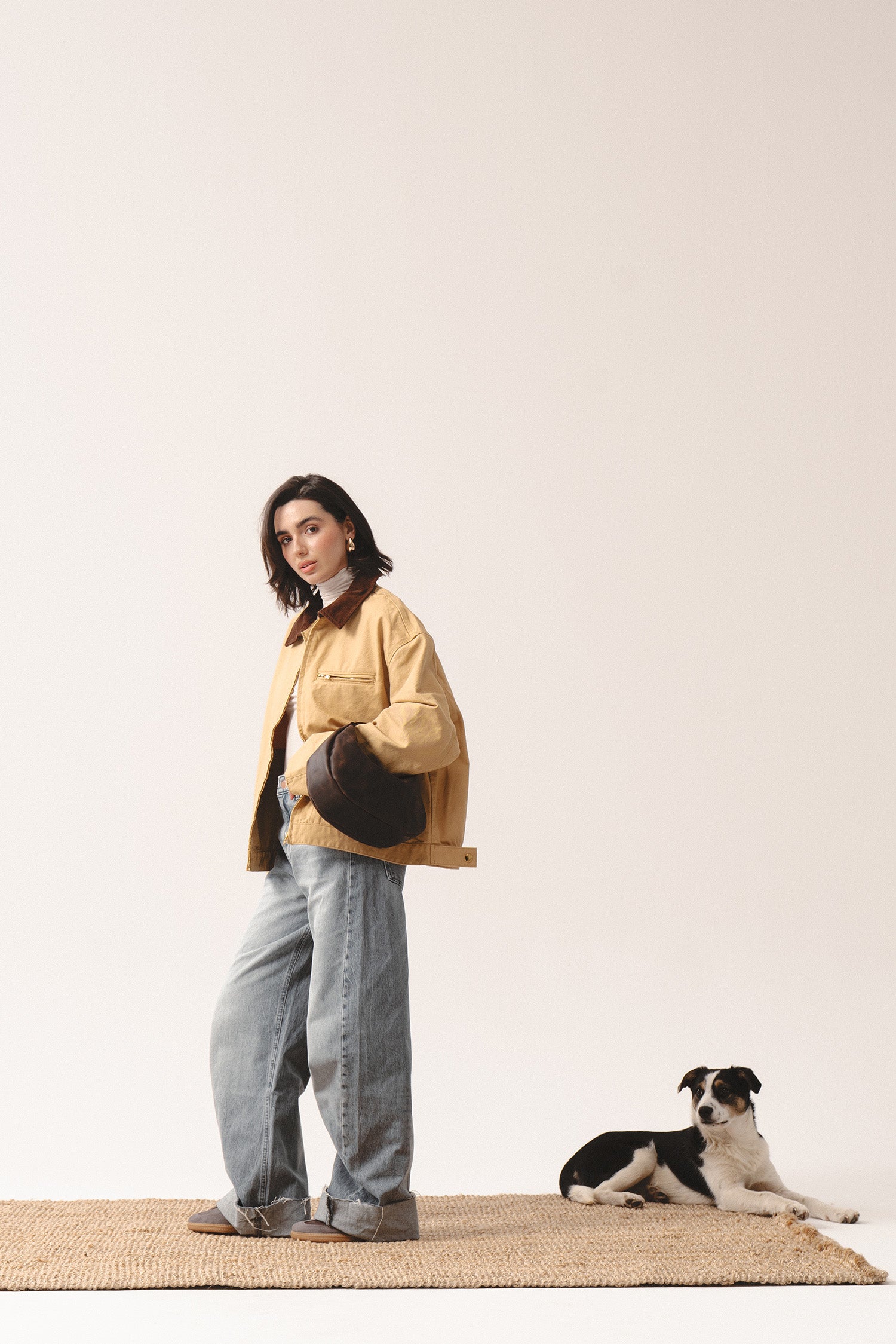 Ciemnobrązowa skórzana torebka Hedera marki Taksuba trzymana pod pachą przez kobietę w beżowej kurtce i dżinsach, z psem w tle